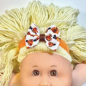 May include: A doll with light yellow yarn hair wearing an orange elastic headband featuring a white ribbon bow. The bow is decorated with a repeating pattern of brown, orange, and yellow turkeys wearing black top hats. The doll has brown eyes.