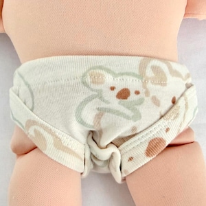 May include: A pair of off-white cotton doll underwear featuring a repeating pattern of light green and tan koala bear illustrations. The fabric has a ribbed texture and the garment is shown on a light-skinned doll torso with visible leg joints.