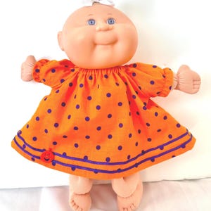 May include: A doll with a round head, blue eyes, and a white hair bow, wearing an orange dress with purple polka dots. The dress features two horizontal purple stripes near the hem and a small, round, orange jack-o'-lantern button on the front.