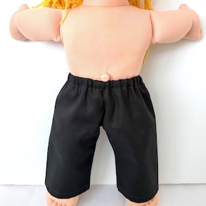 May include: A doll with light skin and yellow yarn hair wearing black pants with an elastic waistband. The doll has a visible belly button and is positioned against a plain white background, showing the torso and legs of the figure.