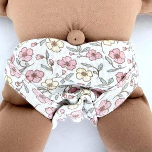 May include: A doll with a tan fabric body and a floral diaper. The diaper features a white background with pink and yellow flowers and green leaves. The doll's arms and legs are visible.