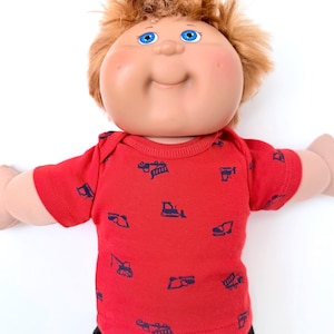 May include: A doll with light skin, bright blue eyes, and messy light brown hair. The doll is wearing a short-sleeved red shirt featuring a repeating pattern of dark blue construction vehicles, including excavators, dump trucks, and bulldozers.