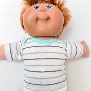 May include: A doll with a large round head, light skin, and short reddish-brown hair. The doll has bright blue eyes and a small smile. It is wearing a short-sleeved shirt with horizontal stripes in shades of blue, green, and white.