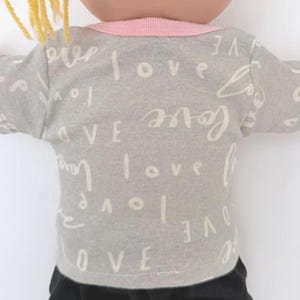 May include: A short-sleeved t-shirt in a light gray color featuring a repeating pattern of the word love written in a white cursive script. The garment has a solid light pink crew neckline and is worn by a doll with yellow yarn hair.