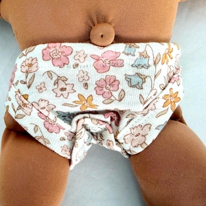 May include: A brown fabric doll wearing white underwear featuring a floral pattern with pink, yellow, and light blue flowers. The doll has a round, centered belly button detail and smooth, tan-colored fabric skin on its torso and legs.