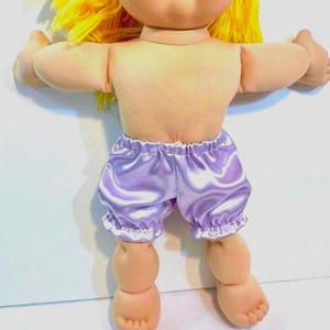 May include: A doll with yellow hair and light skin, wearing lavender satin bloomers with a ruffled hem. The doll's arms are outstretched, and its feet are visible.
