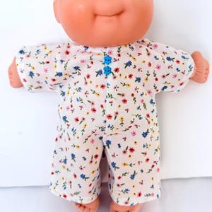 May include: A doll with a round peach-toned head, blue eyes, and a small smile, wearing a white one-piece outfit featuring a small floral pattern in red, yellow, and blue. The garment is fastened at the center front with two small blue flower-shaped buttons.