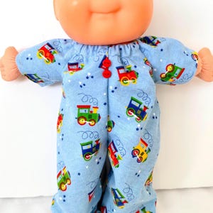 May include: A Cabbage Patch Kid doll wearing light blue pajamas with a colorful train pattern. The pajamas have long sleeves and pants, fastened with two red buttons. The doll has a smiling expression.