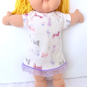 May include: A Cabbage Patch Kid doll with yellow yarn hair and a white dress. The dress features a pattern of pink, gray, and purple animal illustrations, with a lavender lace trim at the hem. The doll has a round face and is posed with arms outstretched.