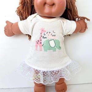 May include: A Cabbage Patch Kid doll with brown hair and brown skin, wearing a white striped dress with a lace hem. The dress features a graphic of a giraffe, zebra, and elephant with the text "L there".