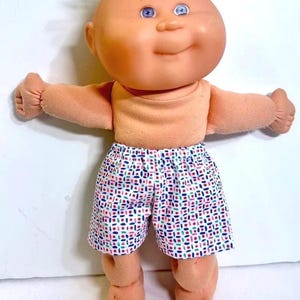 May include: A doll with a round peach-toned head, blue eyes, and light blonde hair. The doll has a soft peach-colored torso and arms, and is wearing white shorts featuring a repeating pattern of small red, blue, and green geometric shapes.