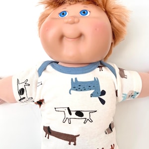 May include: A Cabbage Patch Kid doll wearing a white short-sleeved shirt with a blue neckline. The shirt is patterned with various dog illustrations in black, brown, and blue.