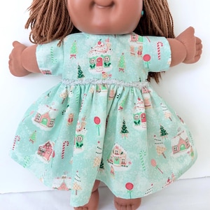 May include: A doll with brown yarn hair wearing a short-sleeved dress made of light green fabric. The dress features a repeating pattern of gingerbread houses, Christmas trees, and candy canes, accented with a silver metallic trim at the waist.