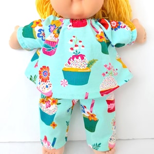 May include: A doll wearing a light blue cupcake-themed outfit. The outfit includes a top and pants, both adorned with colorful cupcake illustrations. The doll has yellow hair and a soft, plush appearance, making it a charming toy.