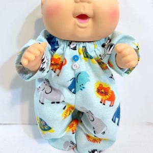 May include: A bald doll with blue eyes and rosy cheeks wearing a light blue long-sleeved romper. The fabric features a colorful print of various animals including lions, elephants, and zebras. The romper has a gathered neckline with two small buttons.