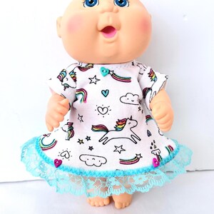 May include: A Cabbage Patch Kid doll wearing a white dress with a unicorn and rainbow pattern. The dress has a blue lace trim. The doll has blue eyes, rosy cheeks, and a surprised expression.