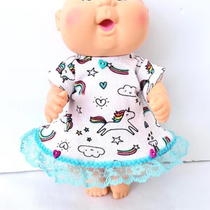 May include: A doll with a round face and pink cheeks wearing a white short-sleeved dress featuring a pattern of unicorns, rainbows, clouds, and stars. The dress has a teal heart-shaped button at the neckline and a teal lace trim at the hem.