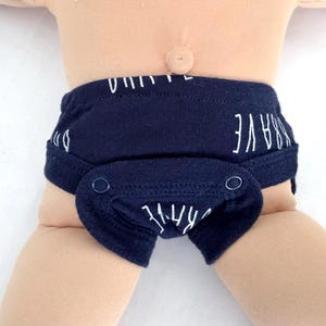 May include: A dark navy blue diaper cover featuring a repeating white text pattern that reads BRAVE. The garment includes two dark blue snap closures on the front panel and is worn on a doll with a light skin tone and a visible belly button.