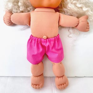 May include: A doll with light peach-colored skin, a soft fabric body, and a prominent belly button. The doll features long, curly, off-white hair and is wearing bright pink elastic-waist shorts. The doll has rounded limbs and defined toes.