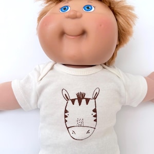 May include: A doll with a round face, bright blue eyes, and messy light brown hair. The doll wears a short-sleeved white shirt featuring a brown line-art illustration of a zebra head on the front, paired with plain black pants.