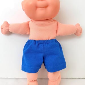 May include: A bald doll with a round peach-colored head, blue eyes, and a small nose. The doll has a peach-colored fabric torso and arms with gathered wrists, and is wearing bright blue fabric shorts with an elastic waistband and bare feet.