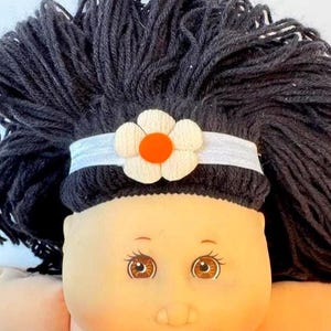 May include: A doll with thick black yarn hair and a light skin tone. The doll features brown eyes and a white elastic headband adorned with a single fabric flower that has white petals and a bright orange circular center, positioned on the forehead.