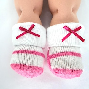 May include: A pair of white ankle socks with pink horizontal stripes on the foot and toe area. Each sock features a folded cuff at the top adorned with a small, dark pink satin ribbon bow centered on the front of the cuff, worn on doll legs.