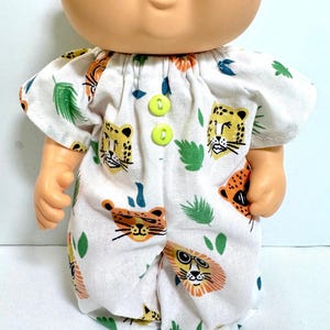 May include: A doll wearing a white romper with a colorful jungle-themed print. The romper features illustrations of lions, leopards, and tropical leaves. The outfit is fastened with two bright green buttons.