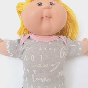 May include: A doll with a round face, light skin, and yellow yarn hair. The doll wears a short-sleeved gray shirt featuring a repeating white script pattern of the word love, accented with a light pink trim around the neckline and shoulders.