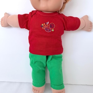 May include: A doll wearing a red short-sleeved shirt and green pants. The shirt has the word "score!" embroidered on it, along with images of a basketball, football, and a star. The doll has red hair and is wearing a red shirt and green pants.