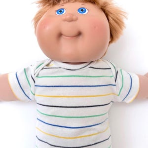 May include: A doll with a round, peach-toned face, bright blue eyes, and messy light brown hair. The doll wears a white short-sleeved shirt featuring horizontal stripes in thin lines of blue, yellow, black, and green, paired with black pants.