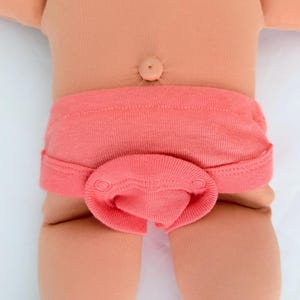 May include: A peach-colored fabric diaper cover with a wide waistband and a front flap secured by two circular snaps. The garment is worn on a doll with a smooth, light-toned torso and a visible belly button, featuring a ribbed texture throughout.