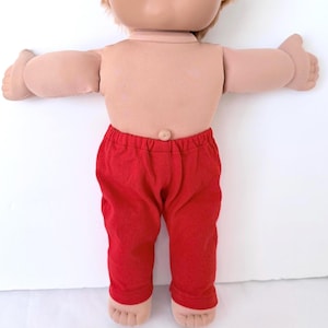 May include: A doll with light skin, short reddish-brown hair, and a soft fabric torso featuring a visible belly button. The doll is wearing solid red pants with an elastic waistband and has outstretched arms and bare feet with defined toes.