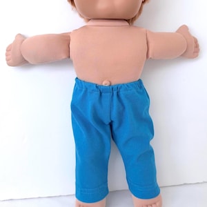 May include: A doll with light skin and short reddish-brown hair wearing bright blue pants. The doll has a soft fabric torso with a visible belly button, outstretched arms, and bare feet. The pants feature an elastic waistband and straight legs.