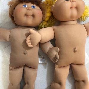May include: Two dolls with soft fabric bodies and vinyl heads. The doll on the left has short brown hair and blue eyes. The doll on the right has yellow yarn hair styled in pigtails. Both dolls feature round faces, small noses, and belly buttons.