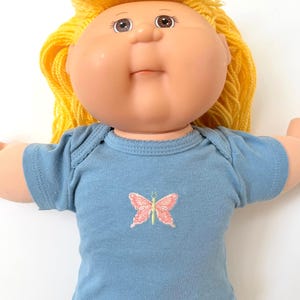 May include: A vintage doll with a round face, blonde yarn hair, and brown eyes. The doll wears a light blue short-sleeved shirt with a pink butterfly design. The doll's arms are outstretched.