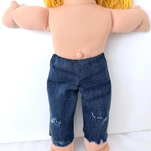 May include: A vintage doll with a soft, flesh-toned body and blonde yarn hair. The doll is wearing dark blue denim jeans with distressed details at the knees and frayed hems. The doll's arms are outstretched.