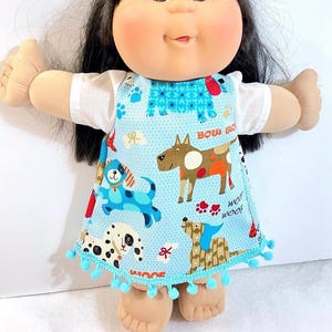 May include: A doll with black hair and a light complexion wearing a light blue dress with a dog pattern. The dress has a white collar and sleeves, and is trimmed with blue pom-poms. The dress features the words "Woof" and "Bow Wow".