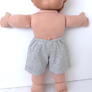 May include: A doll with light skin and reddish-brown hair wearing gray jersey knit shorts. The doll has a rounded face, a small belly button, and outstretched arms. The shorts feature an elastic waistband and a simple hemmed design.