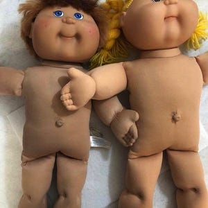 May include: Two dolls with soft fabric bodies and vinyl heads. One doll has brown hair and blue eyes, while the other has yellow yarn hair and brown eyes. Both dolls feature round faces, small noses, and belly buttons, positioned near a ruler.