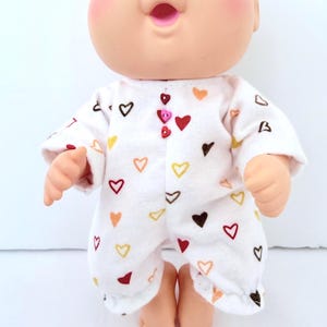 May include: A Cabbage Patch Kid doll wearing a white onesie patterned with colorful hearts. The onesie has a button closure and long sleeves. The doll has blue eyes, rosy cheeks, and a surprised expression.