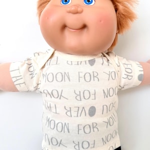 May include: A vintage doll with blue eyes and light orange hair. The doll is wearing a cream-colored onesie with the text "OVER THE MOON FOR YOU" printed in gray. The doll has rosy cheeks and a smiling expression.