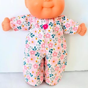 May include: A doll with a peach-colored face and blue eyes wearing a pink floral patterned jumpsuit. The jumpsuit has a pink flower button and features pink flowers, green leaves, and yellow accents.
