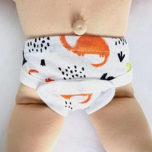May include: A white diaper cover featuring a repeating pattern of orange dinosaurs and black abstract marks. The garment includes a front flap with a snap closure, elasticized leg openings, and a waistband, designed for a doll or small figure.