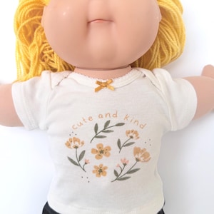 May include: A doll with yellow yarn hair and a cream-colored onesie. The onesie has a floral design and the words "cute and kind" printed on it. The doll has a fair complexion and black pants.