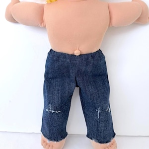 May include: A vintage doll with a soft, flesh-toned body and blonde hair. The doll is wearing dark blue denim pants with distressed details at the knees. The doll's arms are outstretched, and its feet are visible.