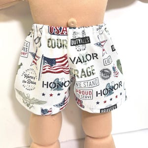 May include: White shorts with a patriotic theme, featuring the American flag, stars, and text like "Honor," "Courage," and "Bravery." The shorts have an elastic waistband and are decorated with various patriotic symbols.