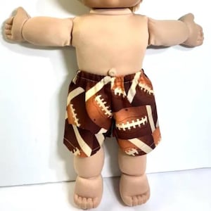 May include: A doll with light skin and light brown hair wearing brown shorts featuring a repeating pattern of brown and white American footballs. The doll has a rounded face, a visible belly button, and is positioned with arms extended outward.