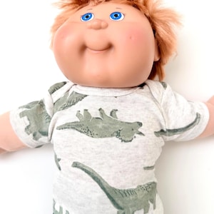 May include: A doll with a round face, bright blue eyes, and messy reddish-brown hair. The doll wears a short-sleeved, light gray heathered shirt featuring a repeating pattern of various green dinosaurs, paired with solid black pants.