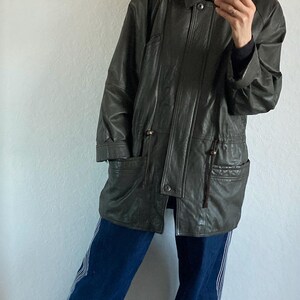 May include: Dark green leather jacket with a fur-trimmed hood. The jacket has a zip and button closure, two front pockets, and a drawstring waist. The person is wearing blue jeans with white stripes and gray shoes.